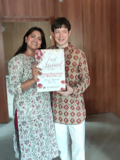 Couple in patterned traditional clothing holding a Just Married placard indoors